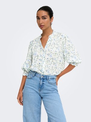 Vrouw met naar achter gestyled haar, gekleed in een witte blouse met bloemenprint en pofmouwen en lichtblauwe high-waisted jeans, staande met één hand op haar heup.