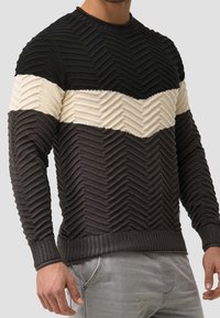 Strickpullover mit Chevron-Muster in Schwarz, Creme und Dunkelgrau. Gerippte Bündchen und Saum, strukturierte Oberfläche, taillierte Passform.