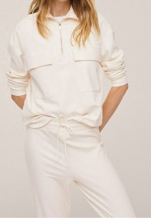 Woman wearing a cream-colored long-sleeve pullover with chest pockets and matching drawstring pants, hands in back pockets.