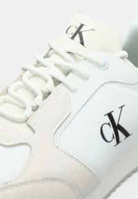 Lichtgekleurde sneaker van glad leer en suede, met een opvallend zwart "cK" logo, platte veters en een gestructureerde zool.