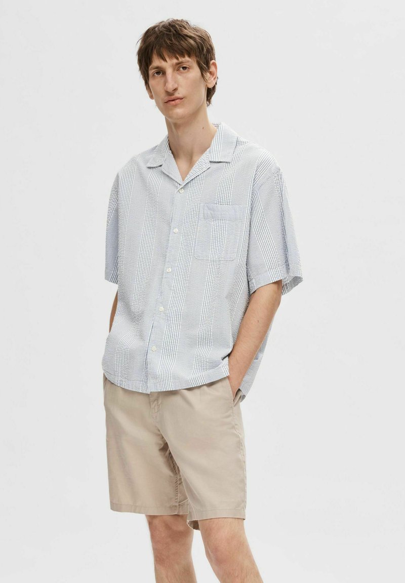 Koszula z krótkim rękawem w jasnoniebieskie i białe pionowe paski, z kieszonką na piersi i zapięciem na guziki, w zestawie z beżowymi szortami.