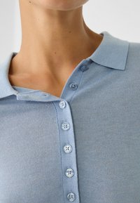 Hellblaues Strick-Polohemd mit Kragen, das über sechs Knopfverschlüsse verfügt und eine glatte Textur aufweist. Der Stoff scheint weich und tailliert zu sein.