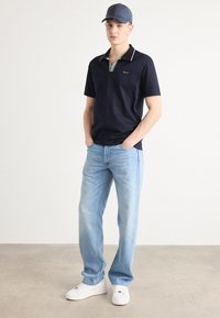 Polo de marina con cuello a rayas, combinado con jeans rectos de color azul claro y zapatillas blancas. El modelo está de pie frente a un fondo neutro.