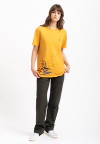 Geel katoenen t-shirt met korte mouwen, met een blauwe grafische afbeelding onderaan en een klein logo op de borst. Gekleed met donkergroene broeken.