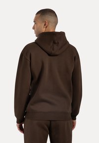 Sudadera marrón de tejido suave, con capucha ajustable y puños acanalados. Costuras visibles y corte holgado.