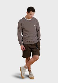 Brown sweatshirt with ribbed cuffs, paired with brown shorts. White high-top sneakers with beige and brown patterns. Model stands with hands in pockets.