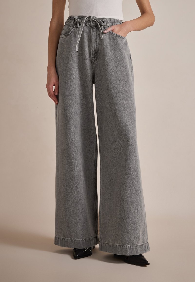 Pantalons en denim gris à jambes larges avec un cordon de serrage à la taille et des poches latérales, présentant une texture douce et une coupe droite et décontractée.