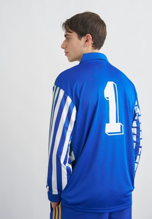 Jaunas vyras mėlynos spalvos sportiniu marškinėliais su baltais skaičiais 1 nugaroje, kurių rankovės dryžuotos mėlynos ir baltos spalvos, stovintis šonu prieš baltą foną.