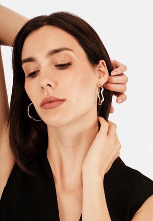 Femme aux cheveux bruns portant un haut noir ajuste des boucles d'oreilles en argent ondulé ornées de petits cristaux sur un fond clair.