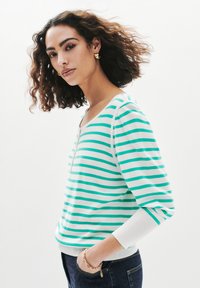 Sweatshirt rayé en blanc et teal, avec un col rond, des manches trois-quarts et un ourlet côtelé. porté avec un jean foncé et des boucles d'oreilles en or.