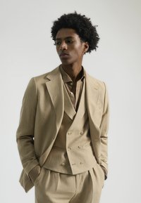 Traje de tres piezas color Beis hecho de tejido texturizado, que incluye un chaleco de doble botonadura, una chaqueta a medida y pantalones con pliegues.