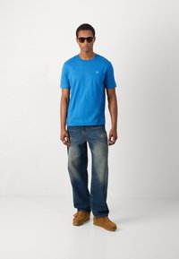 T-shirt de algodão azul com decote redondo, combinado com jeans azuis desbotados e sapatos de camurça bege. A t-shirt apresenta um pequeno logotipo no peito.