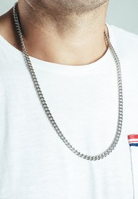 Collana in metallo argento con maglie intrecciate, indossata sopra una maglietta bianca. Presenta una texture liscia e lucida e uno spessore medio.