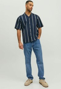 Tamsiai mėlyna trumpomis rankovėmis mygtukinė marškiniai su baltomis vertikaliomis juostelėmis, turintys vieną krūtinės kišenę ir laisvą kirpimą, derinami su šviesiai mėlynomis džinsomis.