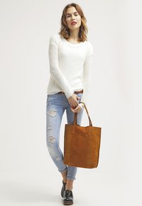 Pull en tricot crème clair à manches longues, associé à un jean bleu clair déchiré et un sac fourre-tout en cuir marron. Chaussures noires lisses.
