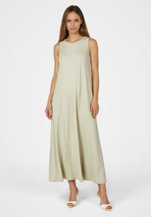 Frau, die ein ärmelloses, bodenlanges beiges Kleid und weiße offene High Heels trägt, steht vor einem schlichten weißen Hintergrund.