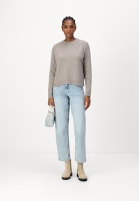 Lichtgrijze gebreide trui met ribbelrand, gecombineerd met lichtblauwe rechttoe-rechtaan jeans en beige enkellaarzen. Een kleine, lichtblauwe handtas wordt vastgehouden.