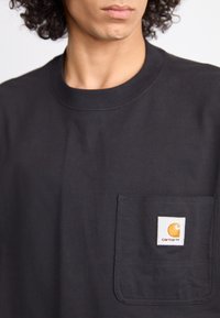 Man wearing black Carhartt T-shirt with a chest pocket featuring a white and yellow logo patch.