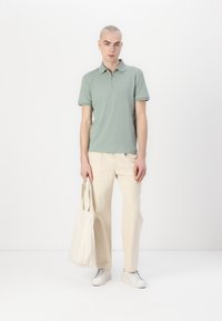 Zelena polo majica z zadrgo na ovratniku, v kombinaciji z svetlo bež hlačami ravne kroja in belimi supergami. Bež torba, ki jo drži v levi roki.
