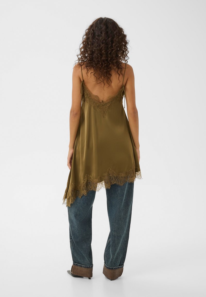 Femme aux cheveux bouclés portant un caraco en satin vert olive bordé de dentelle au-dessus d’un jean bleu ample et de chaussures marron pointues, vue de dos.