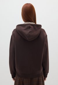 Sudadera marrón con una tela texturizada y un corte relajado, que presenta una capucha y hombros caídos, llevada sobre una camisa de cuadros y una falda de cuadros.