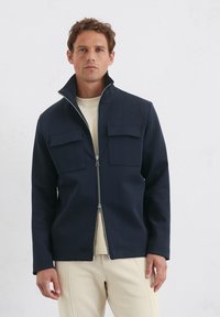 Marineblauwe zip-up jas met hoge kraag, twee borstzakken en een gladde textuur. G gedragen over een crèmekleurig shirt, gepresenteerd tegen een neutrale achtergrond.