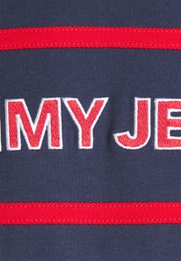 Marinfärgad tyg med röda ränder och broderad vit- och röd text som står "MIN JET." Slät textur med kontrasterande färger och väldefinierade kanter.