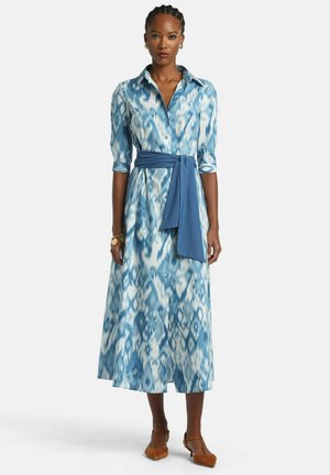 Woman with braided hair wearing a blue and white patterned midi dress with a matching blue waist tie and brown heeled shoes.