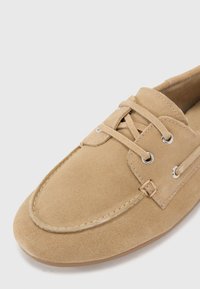Beige Wildlederbootsschuh mit Schnürdesign, silbernen Ösen und abgerundeter Zehenpartie. Weiche Textur und minimale Nähte an den Seiten.