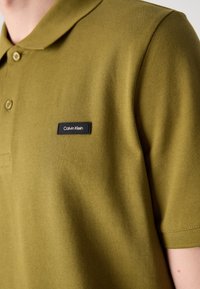 Polo de color verde oliva hecho de tela texturizada, con un cuello clásico, dos botones y un parche de logo negro de Calvin Klein en el pecho.