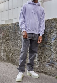 Sweat à capuche lilas avec poche avant, associé à un jean gris. Chaussures avec semelles blanches épaisses et accents translucides. Arrière-plan urbain texturé.