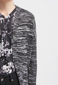 Person som bär en svart och vit blommig blus under en svart, vit och grå stickad cardigan med svarta byxor mot en neutral bakgrund.