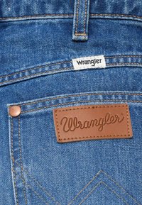 Bolsillo trasero de unos pantalones de mezclilla azul con un parche de cuero marrón de la marca Wrangler y una pequeña etiqueta de tela blanca cosida encima del bolsillo.