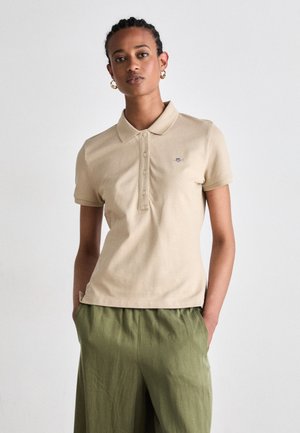 Beige polo skjorte med krage og fire knapper, supplert med en subtil logo. Kombinert med løse grønne bukser.