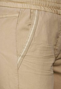 Beiga byxor med ett strukturerat tyg, som har en sidficka och en kontrasterande vit rand längs sömmen. Elastisk midja.