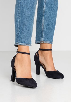 Granatowe szpilki z zamszu z ostrym noskiem, paskiem wokół kostki i wyciętymi bokami. Obcas ma około 10 cm wysokości i gładkie wykończenie.