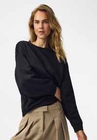 Femme aux longs cheveux blonds ondulés portant un pull noir ample et un pantalon plissé beige sur un fond clair uni.
