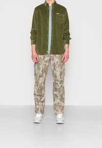 Groene corduroy button-up shirt, lichtblauw T-shirt eronder, camouflage cargobroek en witte sneakers, gepresenteerd tegen een effen achtergrond.