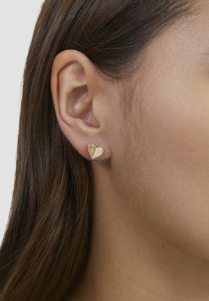 Goldenes Ohrstecker in Herzform mit glatter Oberfläche. Verfügt über eine dezente strukturierte Kante, vor neutralem Hautton platziert. Einfaches, elegantes Design.