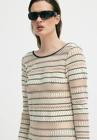 Dlhý rukáv top so vzorovaným sieťovaným dizajnom v krémovej farbe s čiernymi pruhmi. Model s čiernymi slnečnými okuliarmi, neutrálny pozadí.