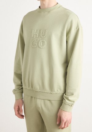 Osoba nosząca jasnozieloną bluzę i pasujące spodnie, bluza ma wypukły napis "HUGO" z przodu.