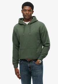 Felpa verde in cotone con tasca frontale, cappuccio regolabile con coulisse e polsini a costine. Abbinata a jeans blu e un orologio.