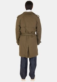 Persona in piedi con le spalle rivolte, che indossa un trench coat verde oliva con cintura, pantaloni blu scuro e scarpe bianche su uno sfondo semplice.