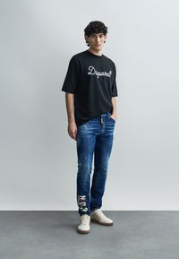 T-shirt preta oversize com texto branco "Dsquared2", jeans azul médio desgastados com salpicos de tinta e gráficos de destaque coloridos, ténis brancos.