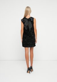 Vestito nero con paillettes, senza maniche, caratterizzato da un grande fiocco in satin nero sul retro e una linea dell'orlo diritta, indossato con tacchi neri.