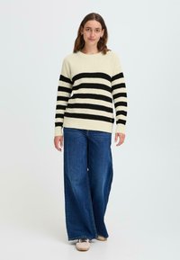 Maglione crema con righe orizzontali nere, texture a coste, scollatura a girocollo, abbinato a jeans blu a gamba larga e sneakers beige.