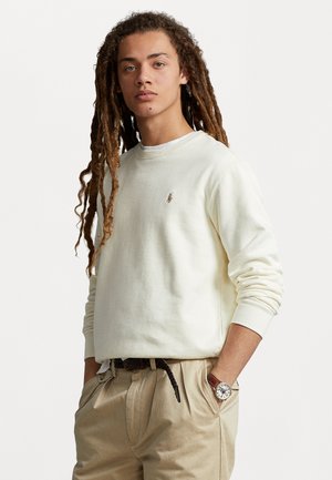 Sudadera crema con cuello redondo, puños acanalados y logo bordado en el pecho. Combinada con pantalones beige y un cinturón marrón. Reloj plateado visible.