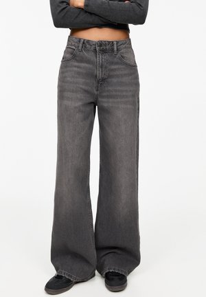 Person trägt hoch taillierte, weit geschnittene, ausgewaschene schwarze Jeans und schwarze Sneakers, Arme verschränkt, graues, gekürztes Langarmshirt, weißer Hintergrund.