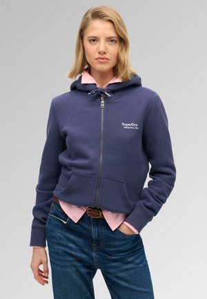 Vrouw die een marineblauwe hoodie met rits draagt over een roze overhemd met kraag en blauwe spijkerbroek, staand tegen een effen lichtgrijze achtergrond.