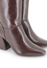 Botas de cuero marrón oscuro hasta la mitad de la pantorrilla con punteras puntiagudas y tacones cuadrados sobre un fondo blanco.